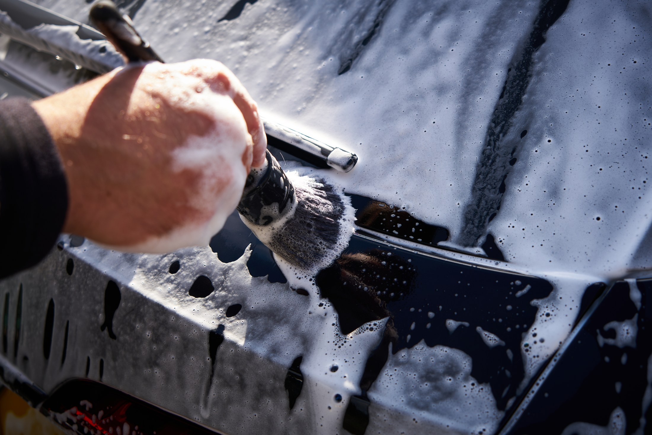 A hand using a detailing brush to clean a soapy car